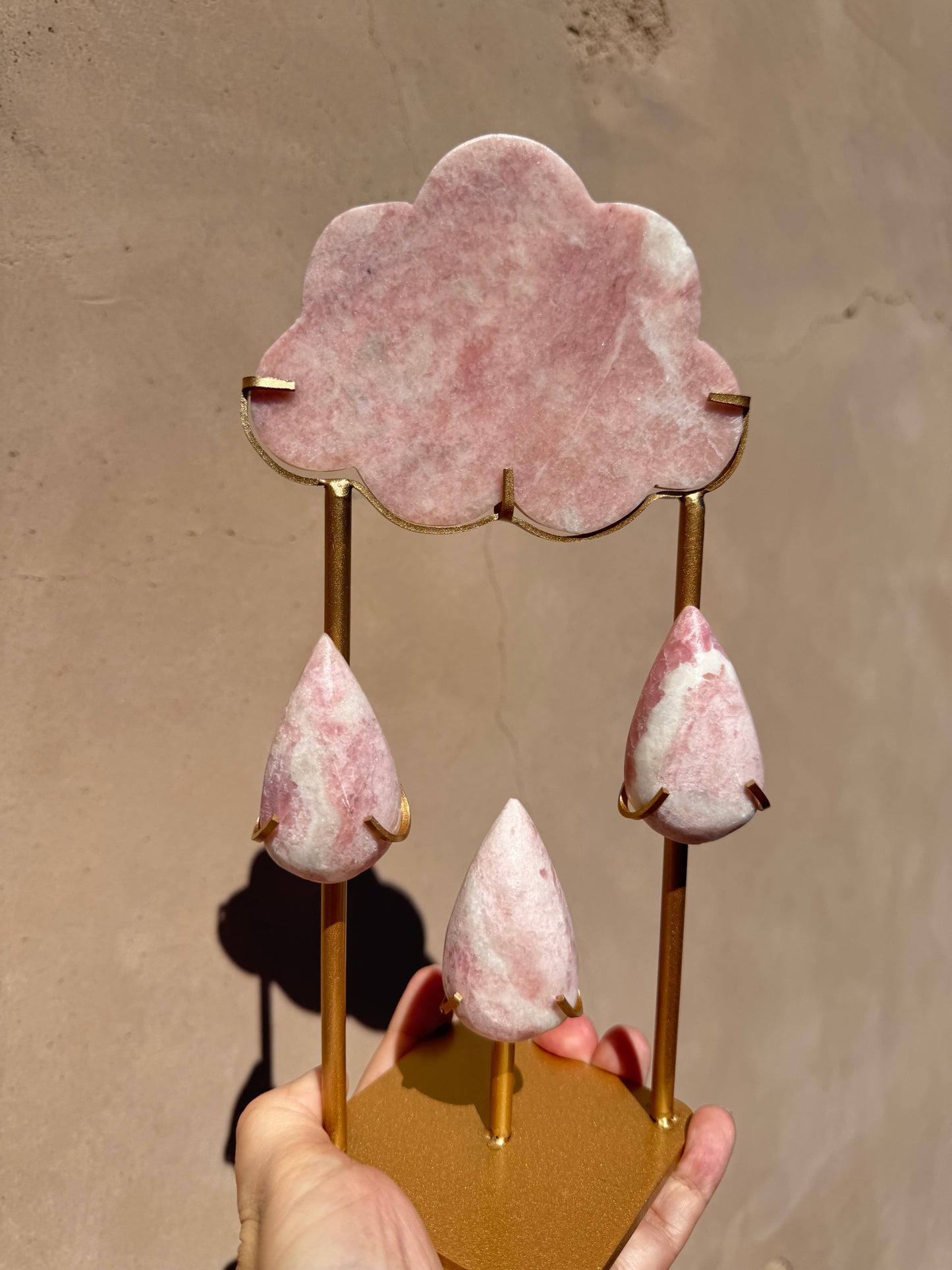 A hand holds the Rain Cloud - Pink Opal, a decorative piece featuring pink opal cloud carvings with three matching teardrop forms suspended below on gold metal rods, evoking the elegant energy of crystal healing.