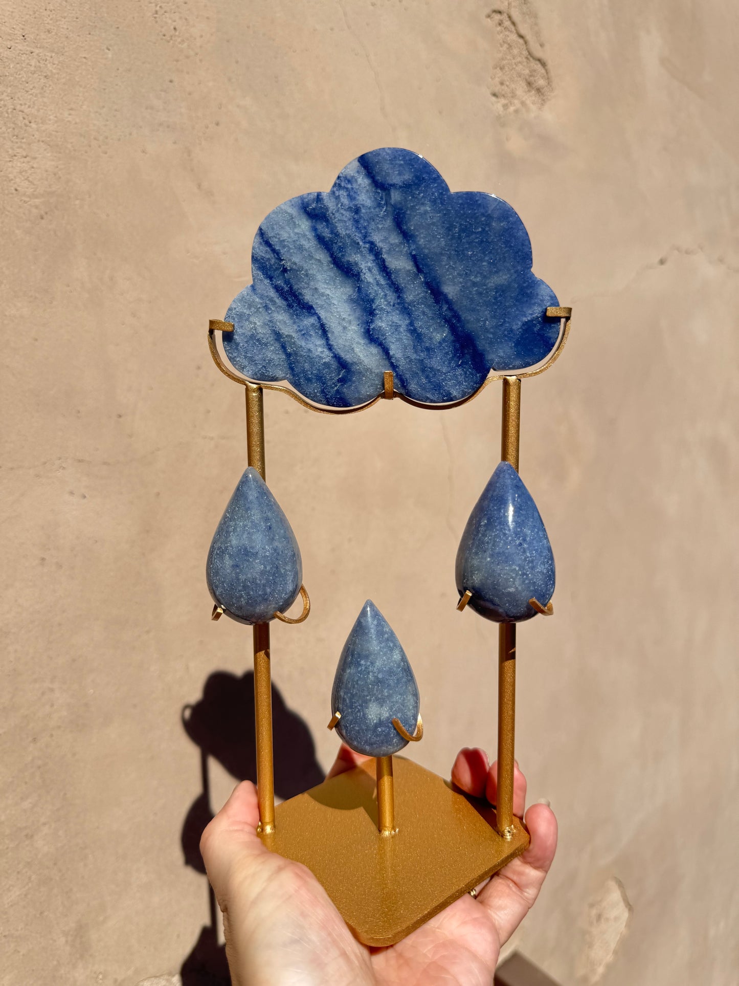 A hand holds the Rain Cloud - Blue Aventurine, showcasing a blue aventurine stone cloud above three blue aventurine raindrops, all skillfully carved from crystal and displayed on a gold-colored stand against a beige background.