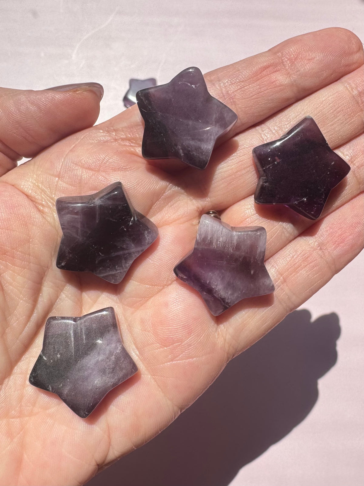 A hand holds five Trapiche Amethyst Star Carvings—small, polished stones in purple and white with smooth surfaces, each uniquely patterned and shown in natural light, prized among healing crystals for their distinct trapiche designs and soothing energy.