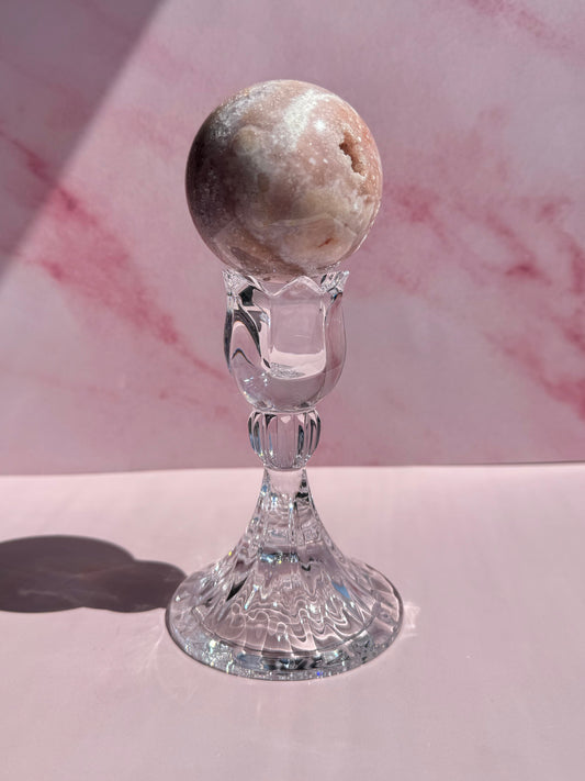 A round, marbled stone sphere is displayed on a clear, ornate Tulip Candle Holder against a pink, marbled background with soft shadows.