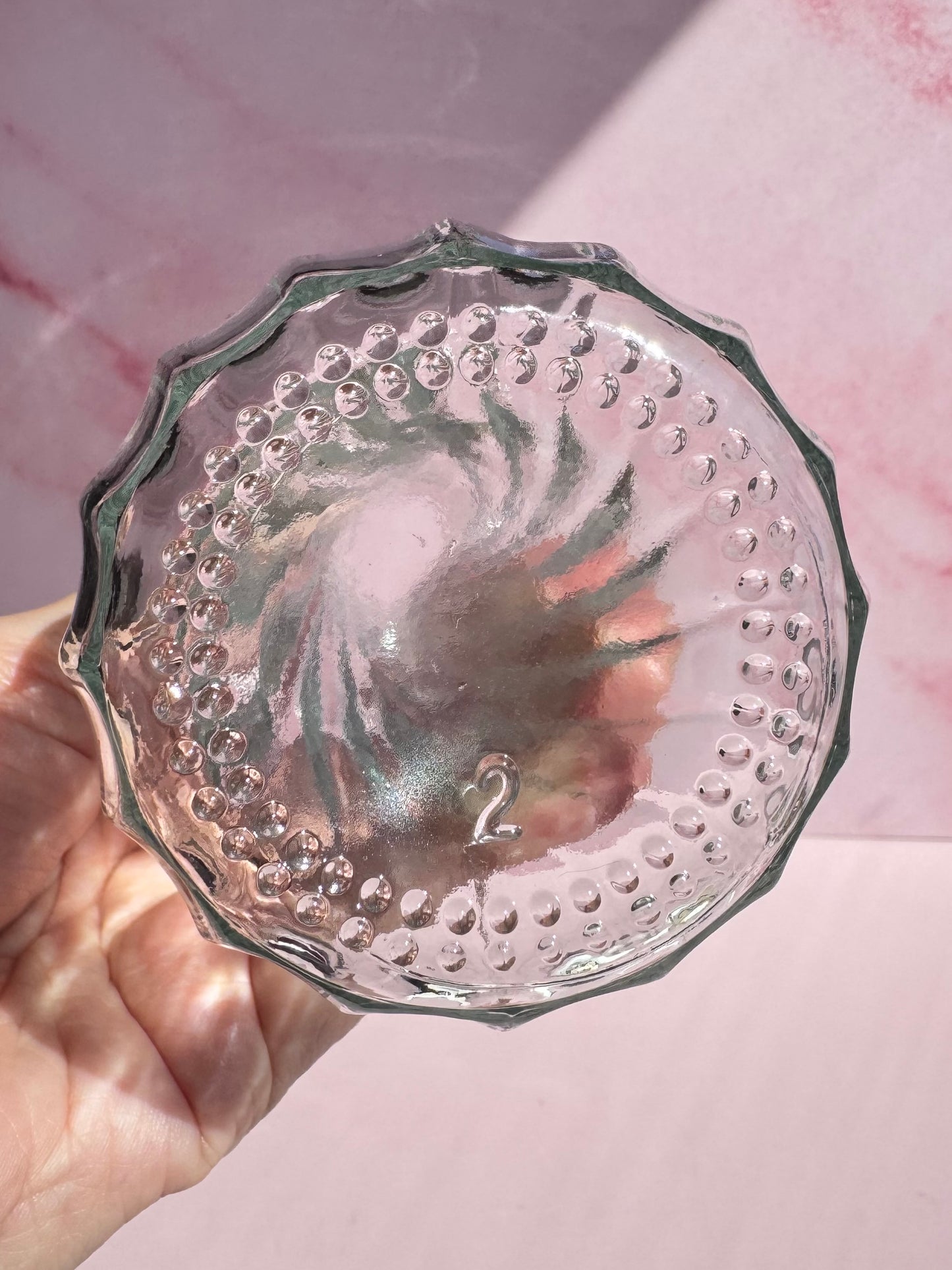 A hand holds the Glass Vase with Spiral Pattern, featuring a clear glass design with a textured, swirled bottom and the number 2 embossed at its center, against a pink background.