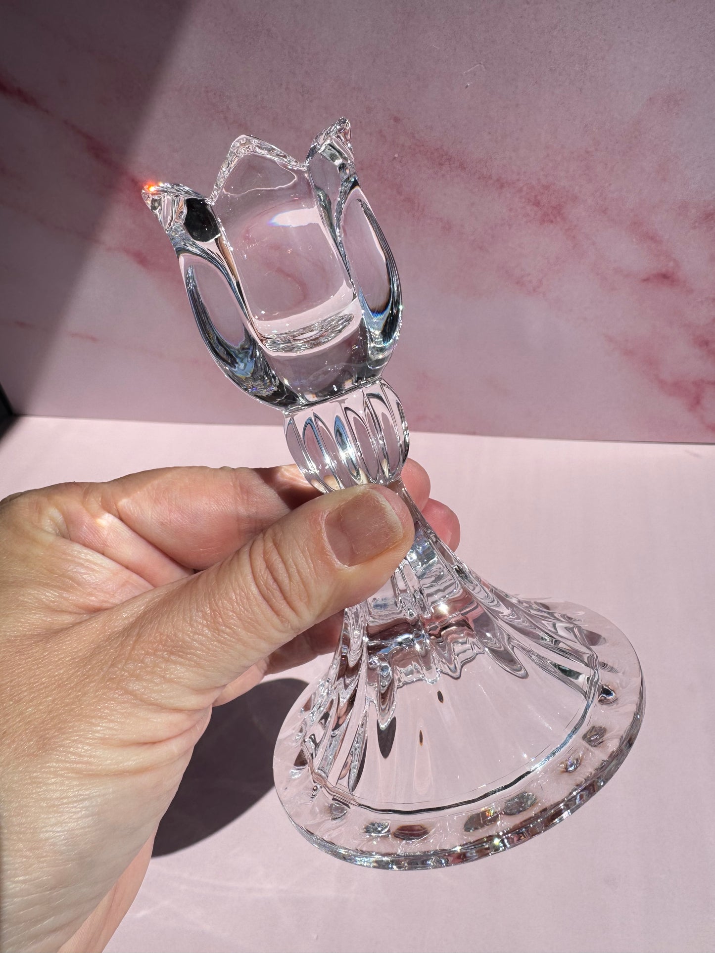 A hand holds the Tulip Candle Holder, a crystal piece with a refined, clear design and distinctive tulip-shaped top, set against a pink marbled background. Inspired by 1990s Mikasa lead crystal pieces from Slovenia, this candle holder adds vintage charm to any setting.