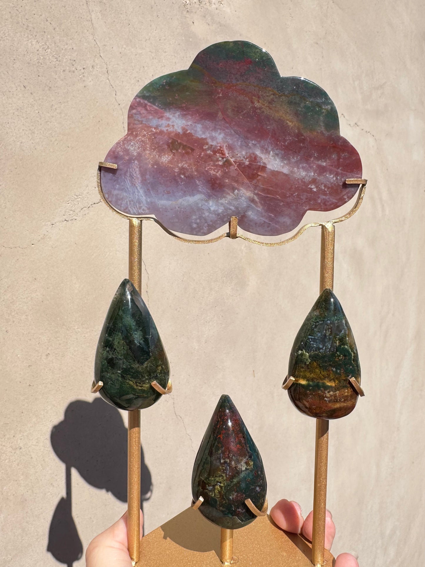 A hand holds the Rain Cloud - Sea Jasper, featuring a sea jasper cloud carving atop a gold metal stand with three teardrop-shaped stones suspended below, casting a gentle shadow on a beige background—perfect as meditation decor or for infusing your space with crystal healing energy.