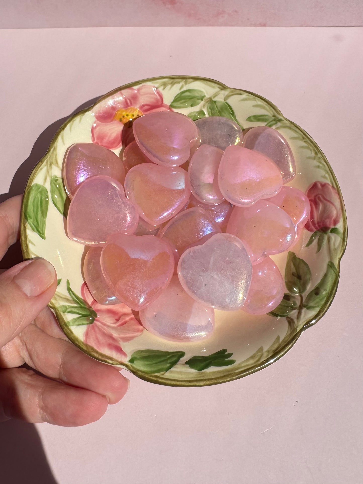 A hand holds a Desert Rose Berry Bowl filled with shiny, pink, heart-shaped objects against a light pink background.