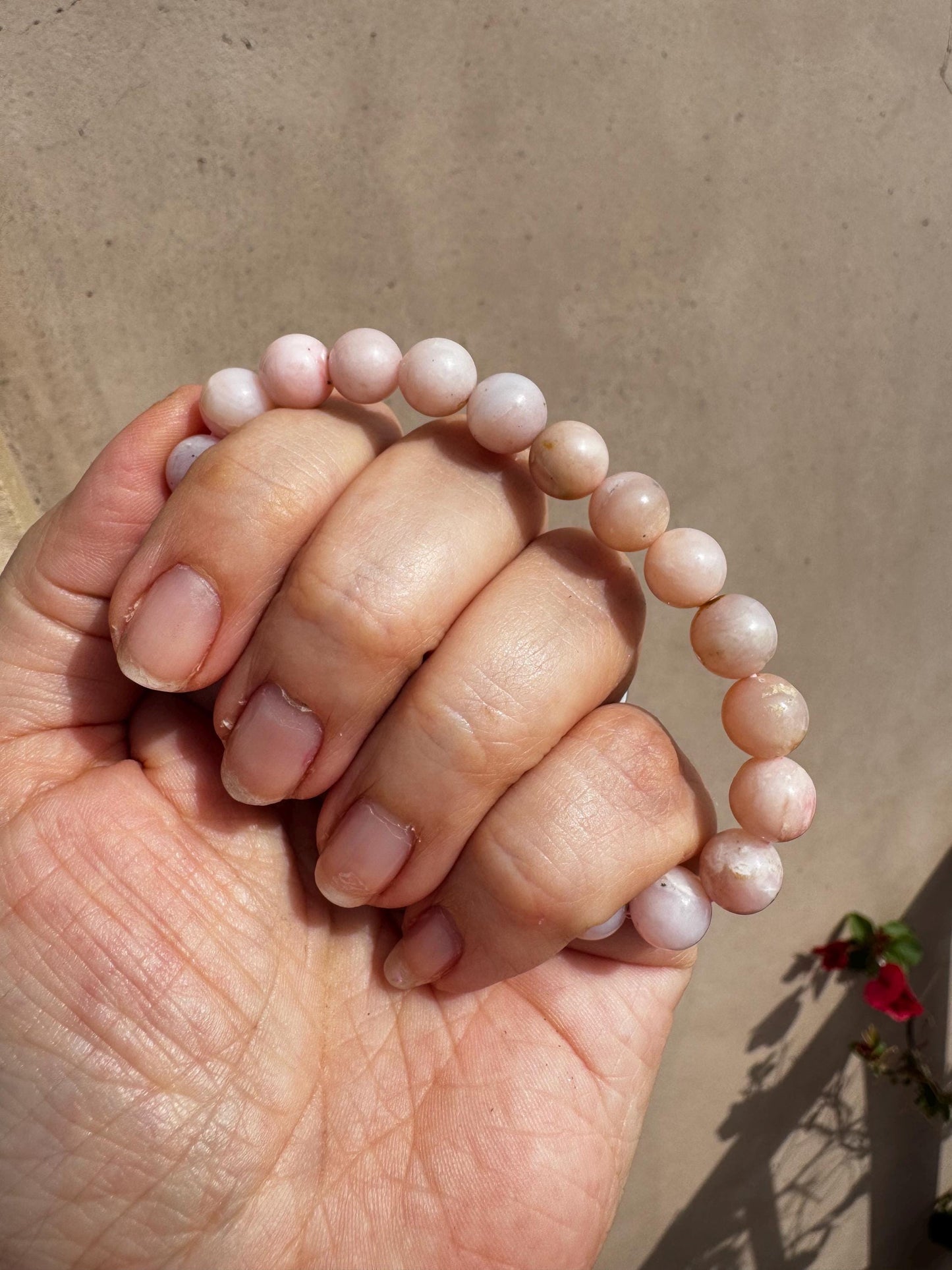 A hand displays a Pink Opalite Bracelet (8mm stretchy, man-made crystal, 6" size) with round light pink beads against a beige background and a small plant—ideal for crystal healing and emotional balance.