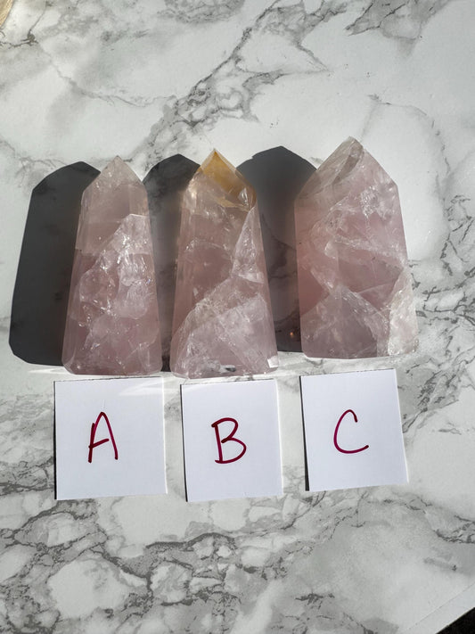 Three Nice Quality Rose Quartz Obelisk Towers from Brazil (3.6"–3.8") labeled A, B, and C rest on marble, each with a card in front—choose your favorite rose quartz obelisk to showcase its natural beauty.