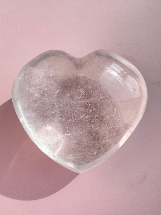 The Clear Quartz Heart Carving - Very High Clarity Crystal rests on a pale pink surface, casting a soft shadow and radiating the calming energy associated with crystal healing.