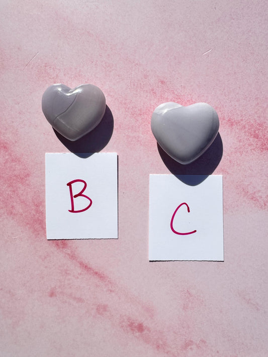 Two Lavender Fluorite Heart Carvings from Mexico, labeled "B" and "C," rest on a pink surface, each above a white card with a red letter.