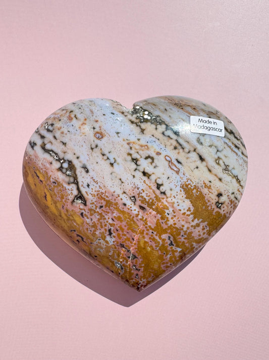 An Ocean Jasper Heart from Madagascar - Item C, featuring white, beige, and brown patterns, rests on a pink surface. A small "Made in Madagascar" sticker is attached.