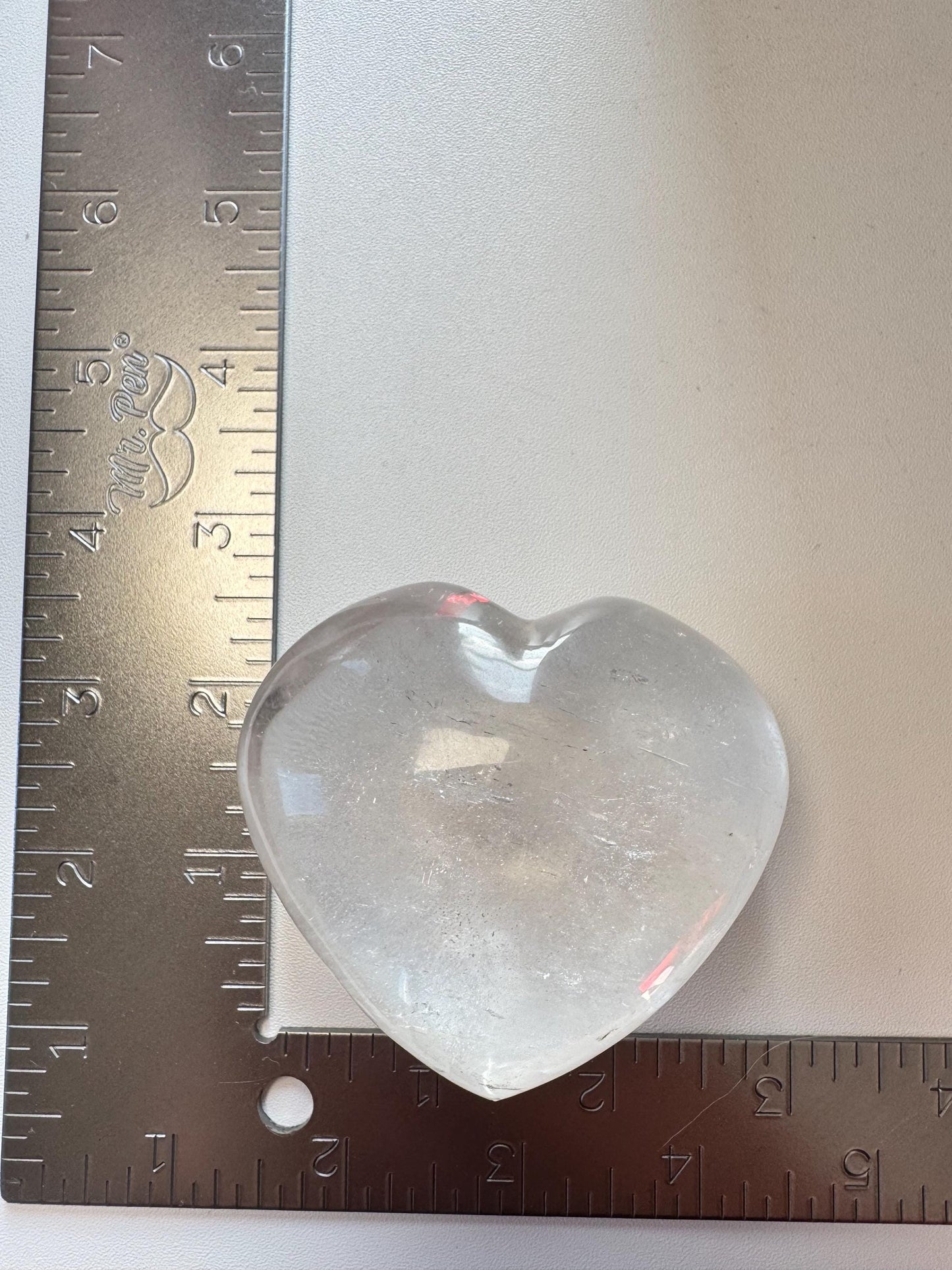 The Clear Quartz Heart Carving - Very High Clarity Crystal is shown next to two metal rulers in an L shape, measuring about 2.5 inches wide and tall—ideal for crystal healing or as a collectible quartz carving.