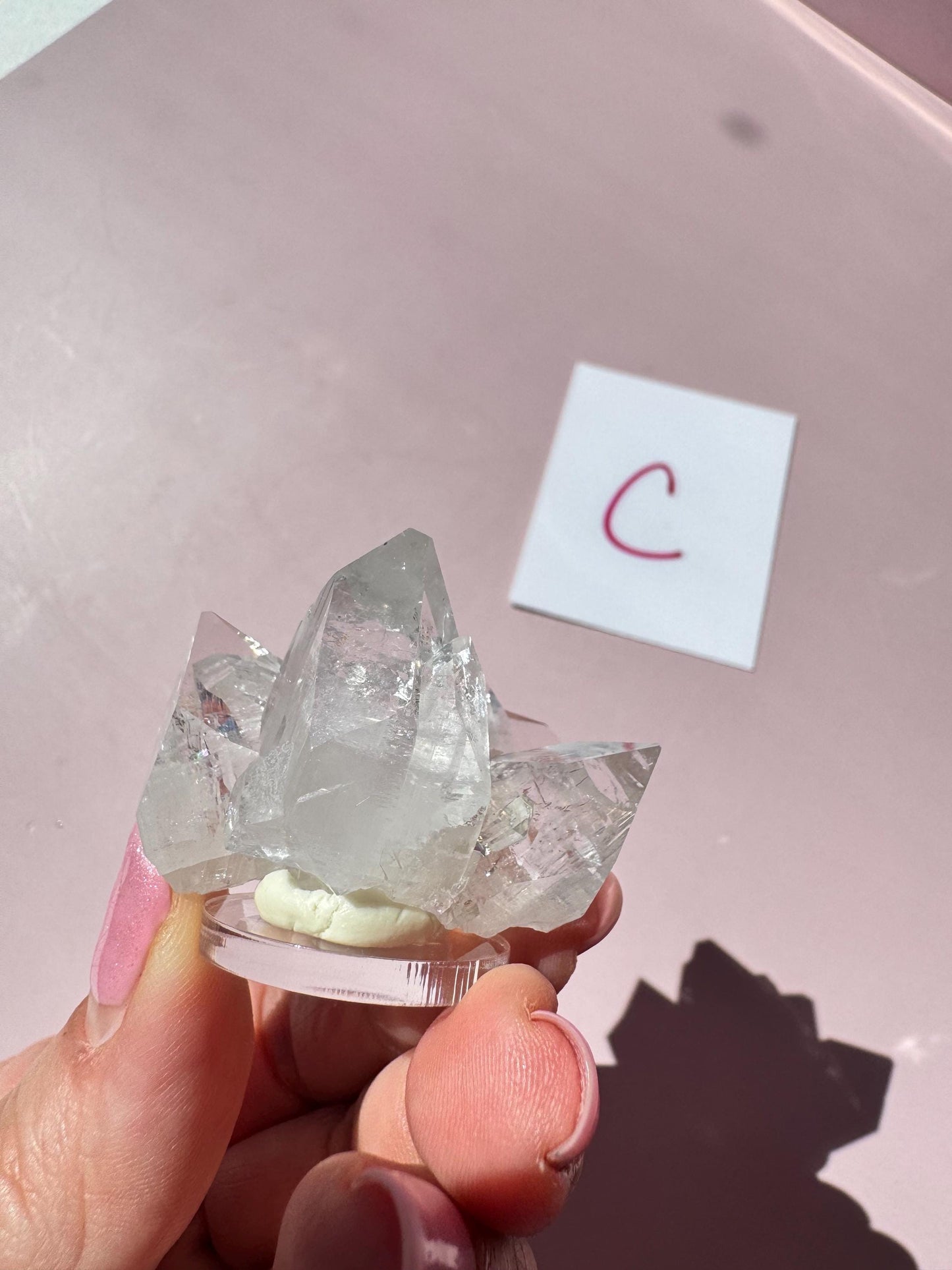 A hand displays the Apophyllite Cluster from India - ITEM C on a round base, with a white card marked “C” in the background atop a pink surface, highlighting the product’s association with spiritual clarity.