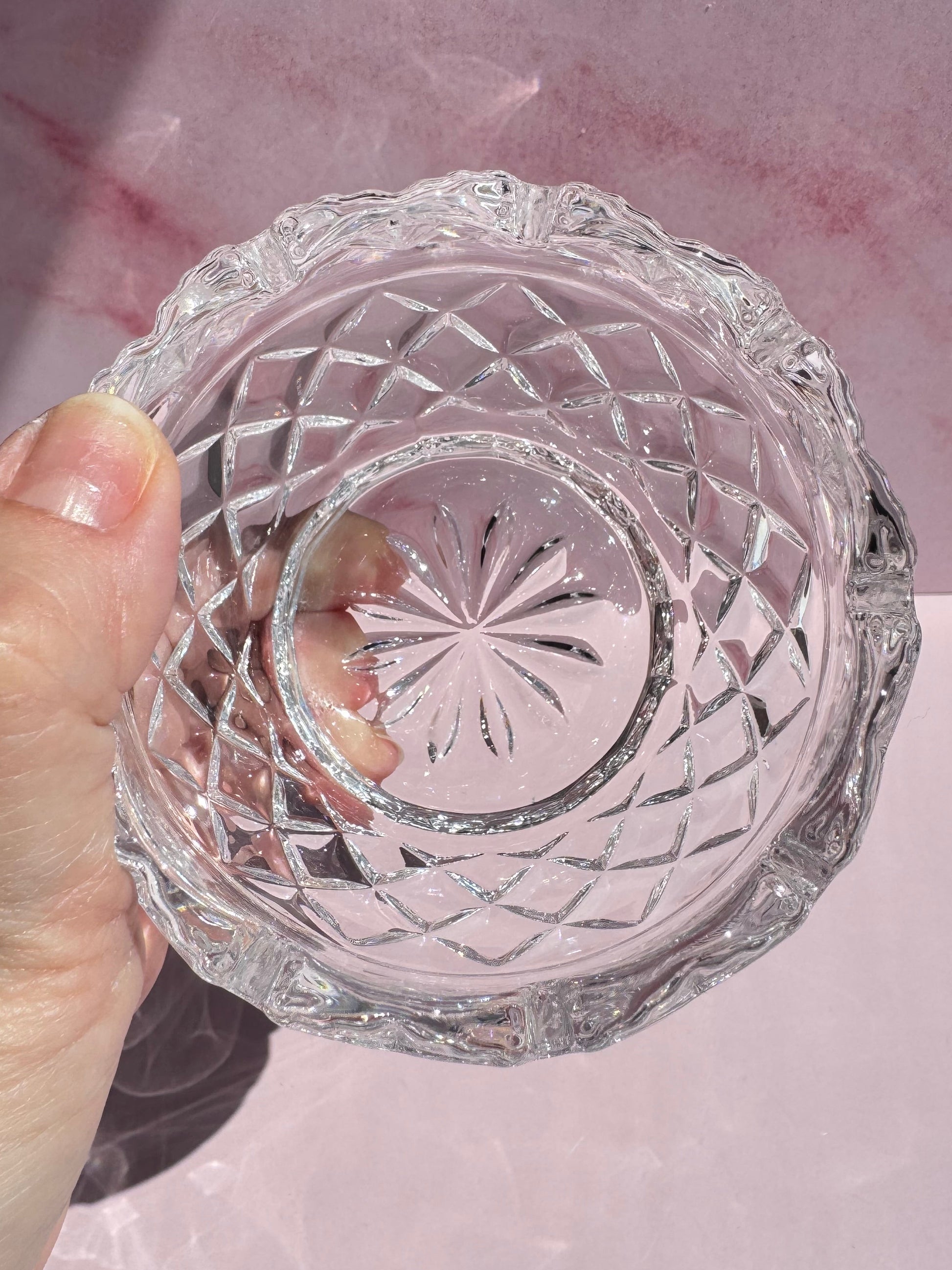 A hand holding the Cut Crystal Mini Glass Bowl, featuring clear glass with scalloped edges and a starburst pattern on the bottom, against a pink background.