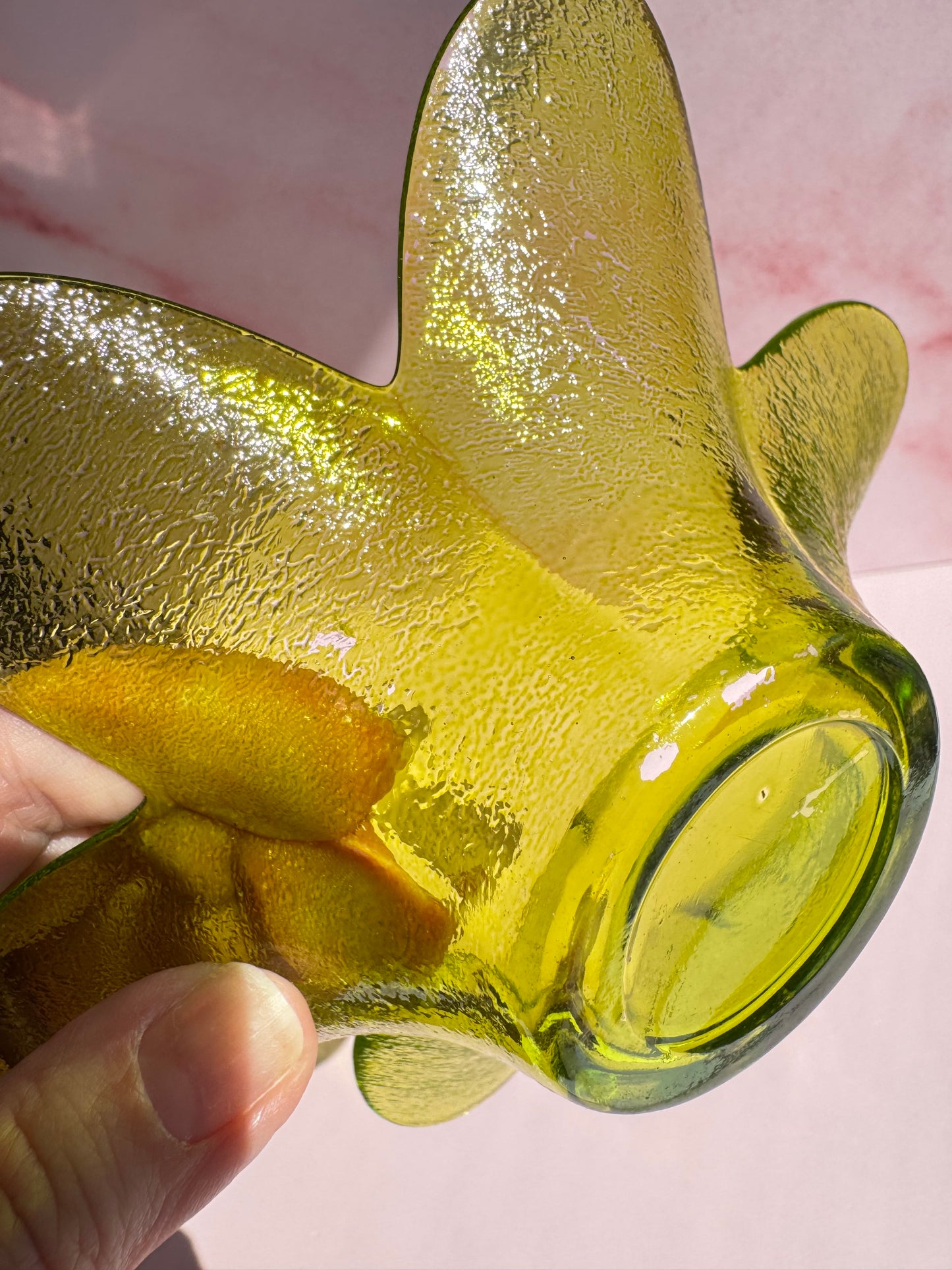 A hand holds the Green Flower Glass Bowl (Imperfect), its textured yellow-green glass and curved edges highlighted against a pale pink background in sunlight.