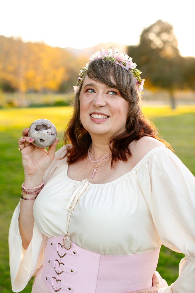 A woman wearing a flower crown and off-the-shoulder dress stands outdoors, smiling and holding a donut in her right hand.