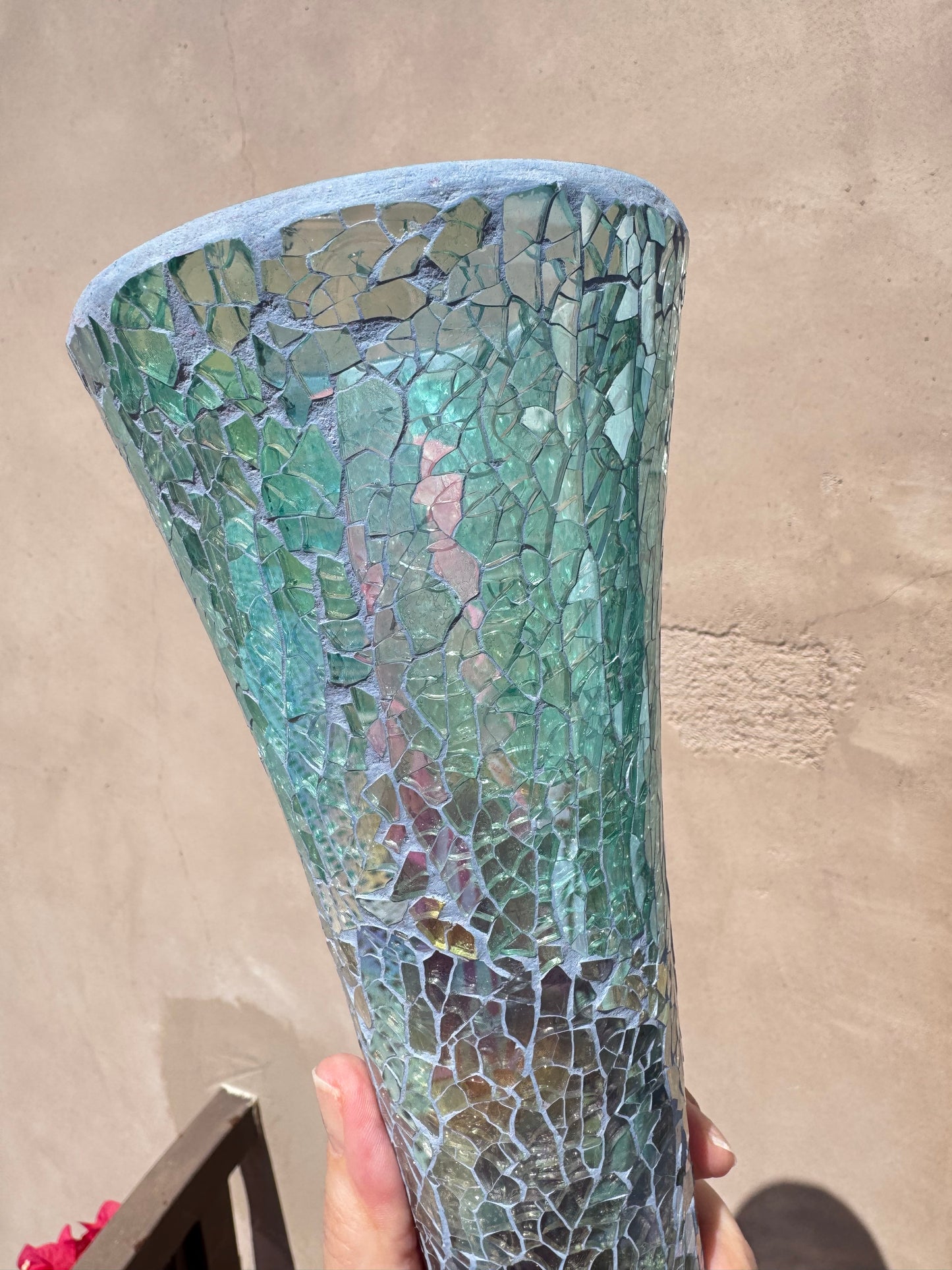 A hand holds the Iridescent Blue Mosaic Crackle Glass Vase—a tall vessel with a greenish-blue hue and crackle mosaic pattern—against a beige wall in bright sunlight.