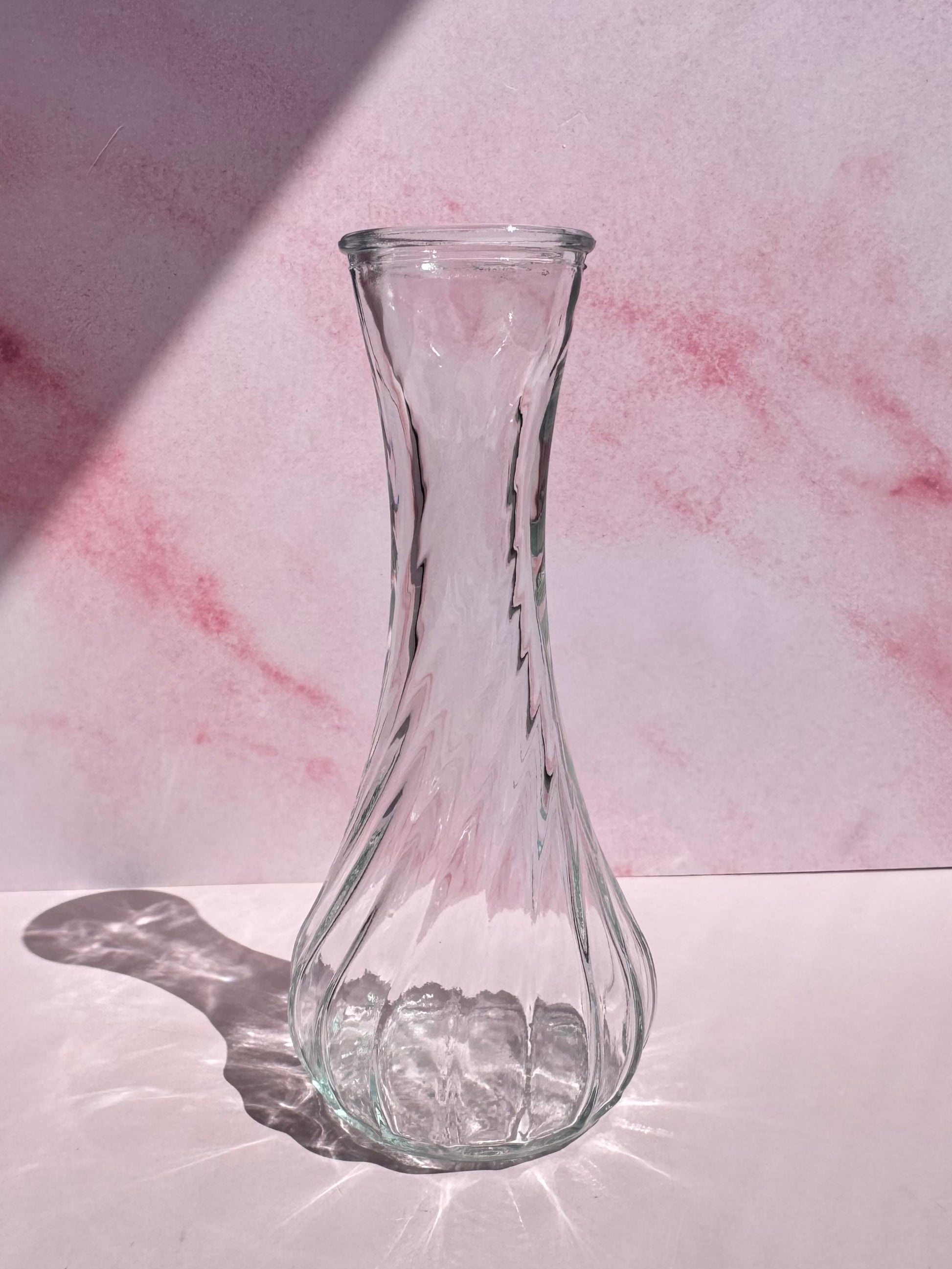 The Glass Vase with Spiral Pattern sits on a white surface against a pink marbled background, casting a shadow to the left. This elegant small vase is perfect for showcasing single blooms or minimalistic arrangements.