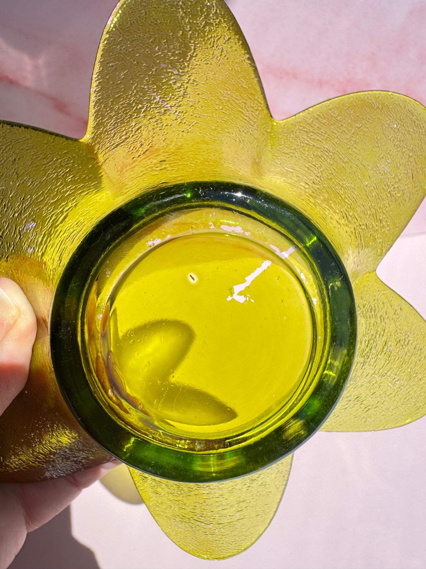 A close-up of the bottom of the Green Flower Glass Bowl (Imperfect), held against a pink surface with sunlight reflecting off its yellow-green glass.