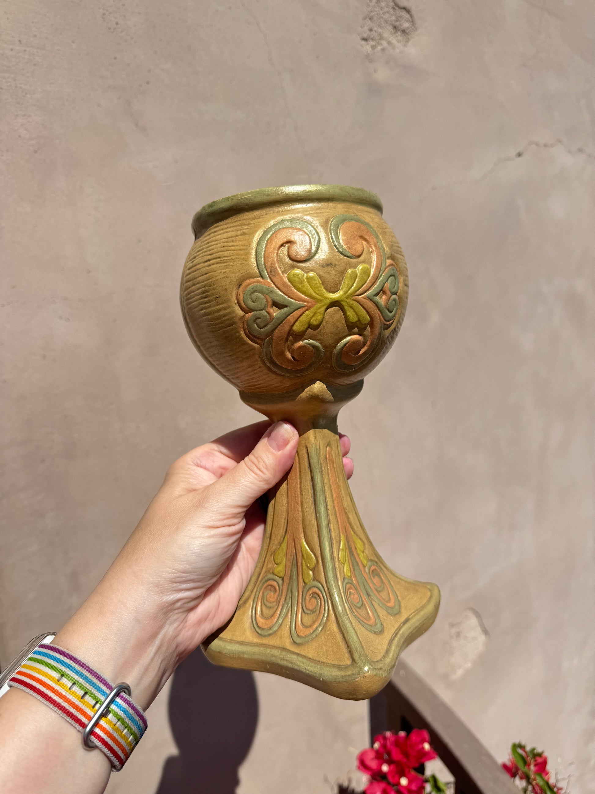 A hand holds the Ceramichrome Chalice Vase, an ornate tan and green ceramic piece with decorative swirl patterns reminiscent of a California vintage chalice, against a beige wall in sunlight.