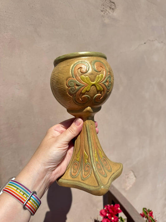 A hand holds the Ceramichrome Chalice Vase, an ornate tan and green ceramic piece with decorative swirl patterns reminiscent of a California vintage chalice, against a beige wall in sunlight.