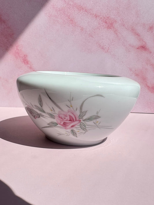 The Porcelain Floral Sugar Bowl features a white ceramic design adorned with a pink rose and green leaves, evoking the charm of vintage Japanese porcelain, and is beautifully displayed on a pink surface with a pink marbled background.