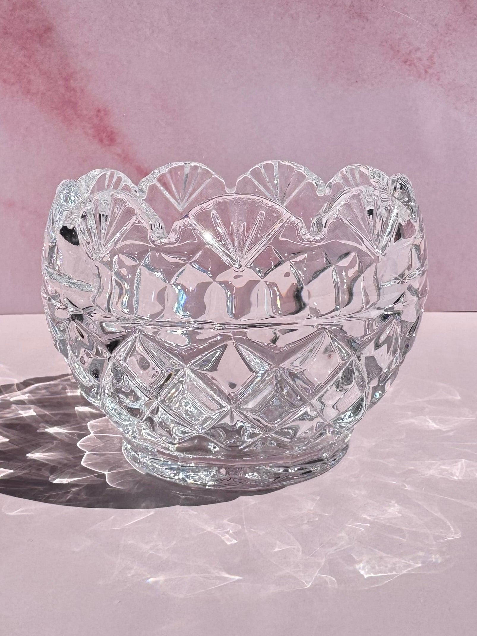 The Cut Crystal Mini Glass Bowl, featuring clear glass with scalloped edges, sits on a light pink surface and casts diamond-patterned shadows and intricate reflections.