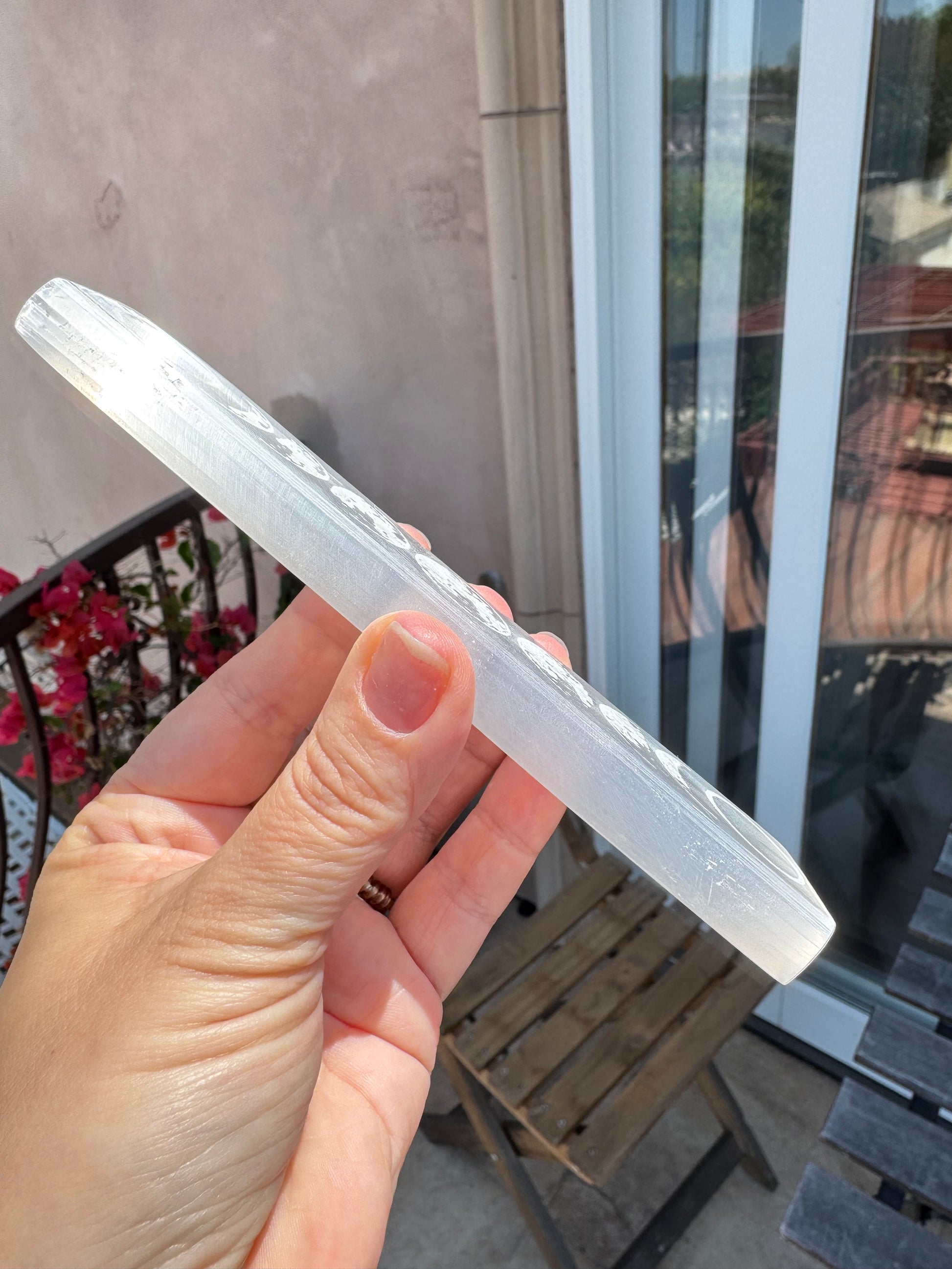A hand holding the Satin Spar Moon Phases Charging Bar—an elegant, translucent white wand-shaped crystal—outside near a balcony, with flowers and a wooden chair visible, perfect for your moon phase crystal rituals.
