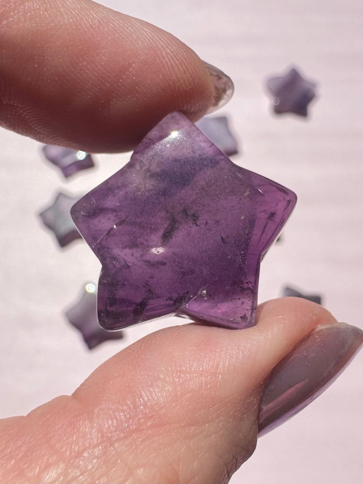 A hand holds a Trapiche Amethyst Star Carving, its polished purple surface expertly shaped into a star, with similar healing crystals softly blurred in the background on a light surface.