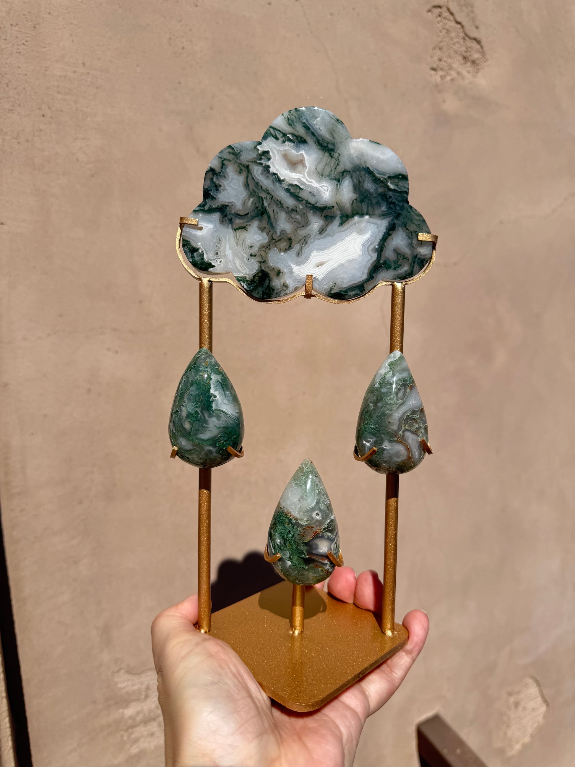 A hand holds the Rain Cloud - Moss Agate, a gold-colored metal stand shaped like a cloud and adorned with three hanging moss agate teardrops, set against a plain tan wall—a perfect accent for those who appreciate crystal healing décor.