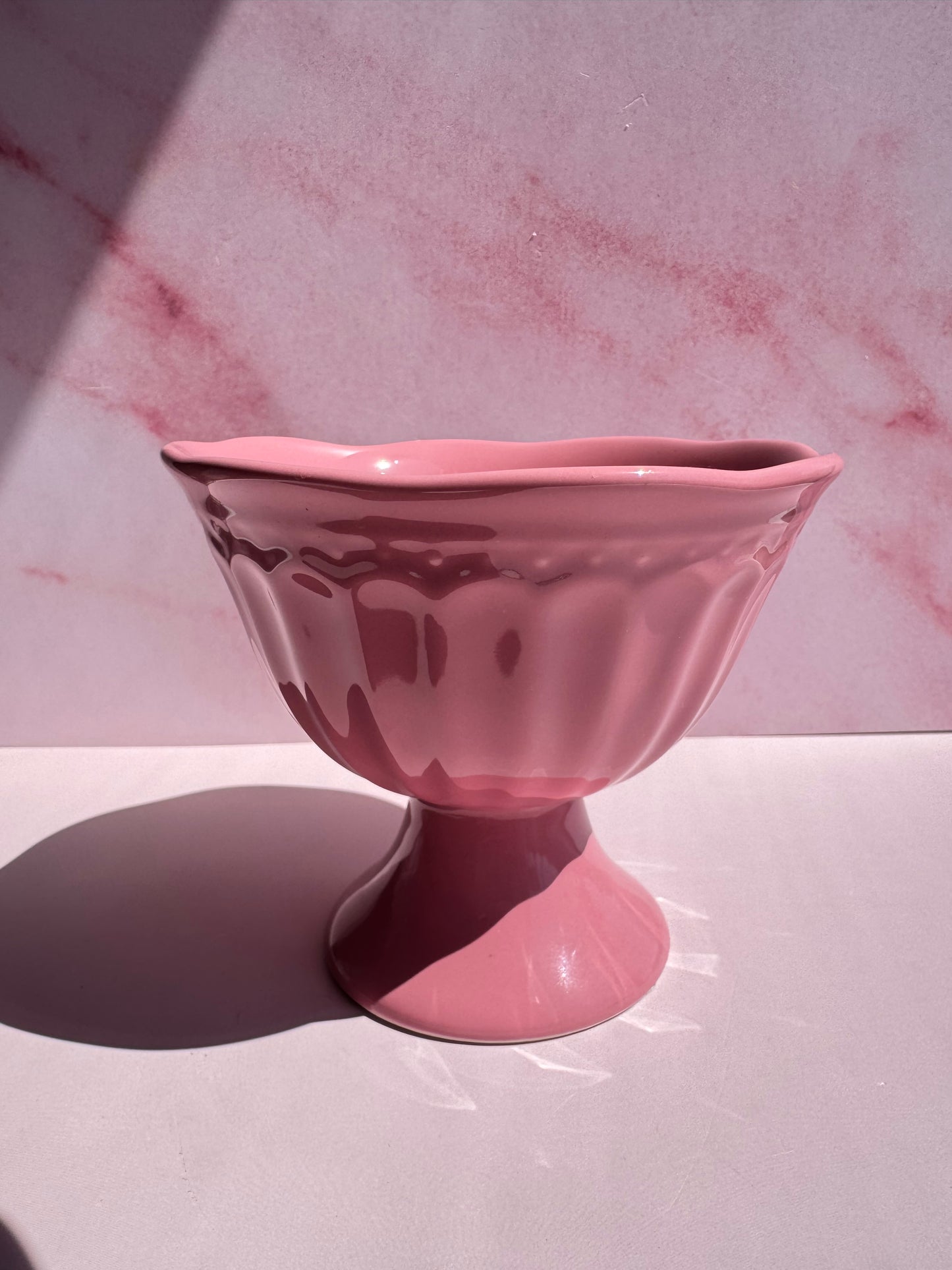 A Pink Ceramic Raised Bowl with a pedestal base sits on a pink surface against a pink marble-patterned background. This charming bowl features a scalloped rim, adding an elegant touch to any table setting.