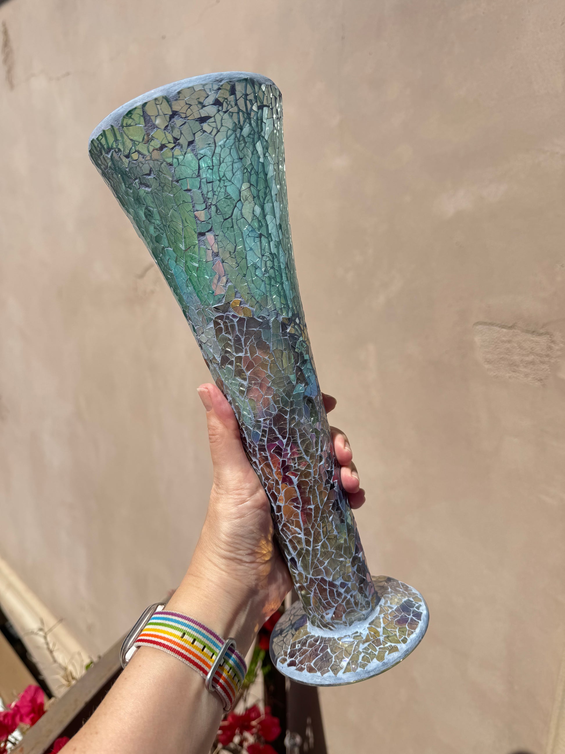 A hand holds an Iridescent Blue Mosaic Crackle Glass Vase, its tall flared shape and blue-green crackled patterns displayed against a plain beige wall.