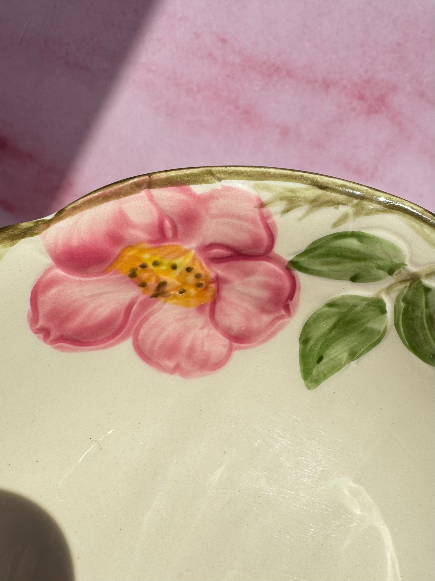 Close-up of a Desert Rose Berry Bowl, featuring a hand-painted pink flower with green leaves and a yellow center on a cream background, set atop a pink surface.