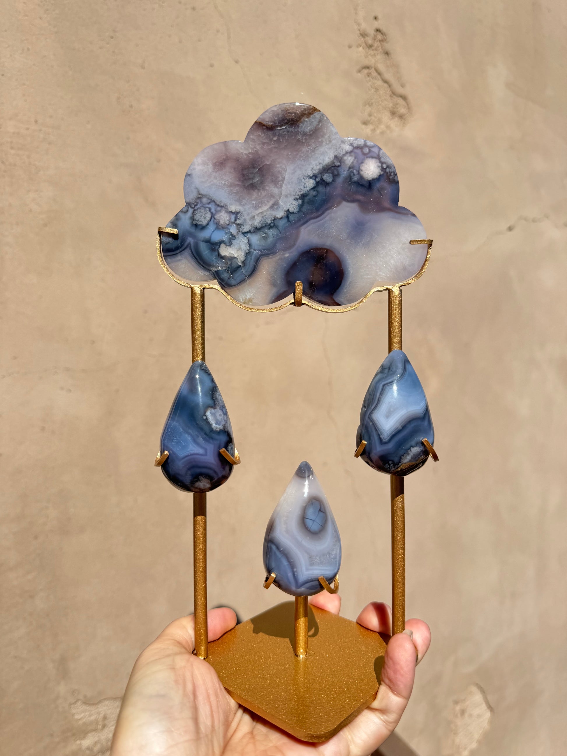 A hand holds the Rain Cloud - Blue Agate, a gold metal stand featuring three teardrop-shaped polished blue chalcedony agate stones and a larger cloud carving on top, set against a plain beige background.