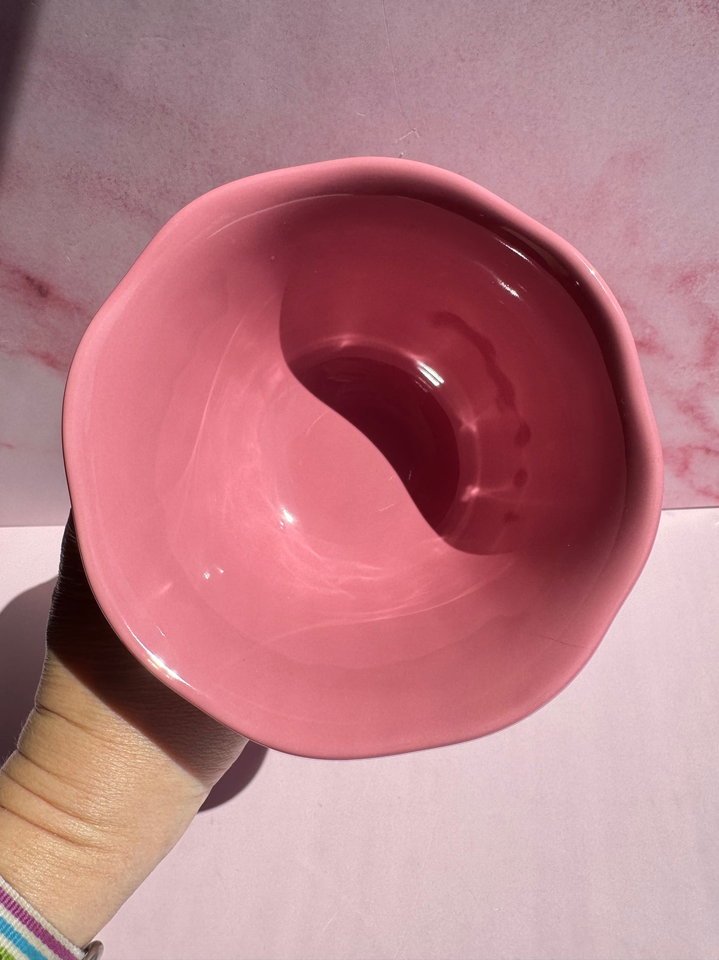 A hand holds the Pink Ceramic Raised Bowl, featuring wavy scallop edges, against a pink marbled background.