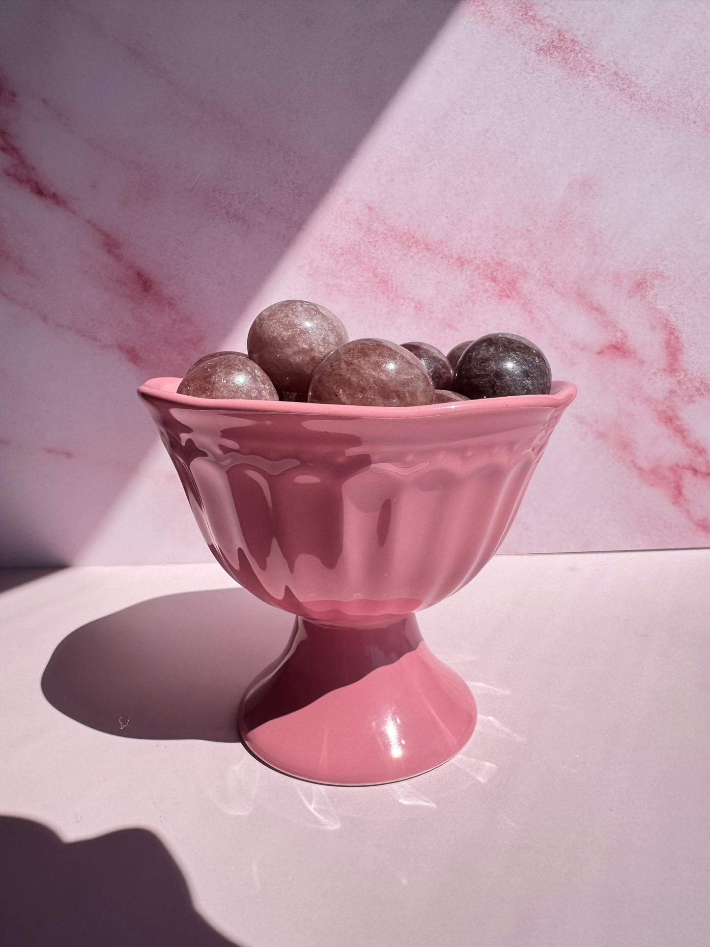 A Pink Ceramic Raised Bowl holds several round, shiny marbles or stones, set against a pink marble-patterned background with sunlight casting shadows.