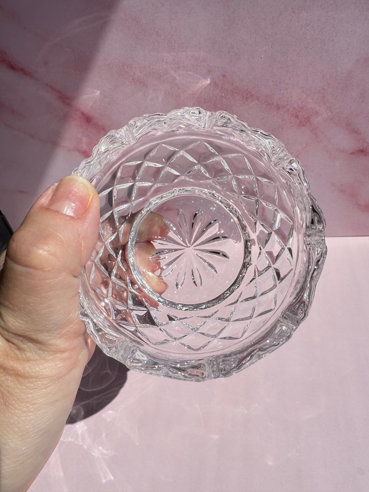 A hand holds the Floral Crystal Bowl— a small, clear, cut glass bowl featuring a starburst pattern on the bottom and an etched delicate flower design along the rim—against a pink background.
