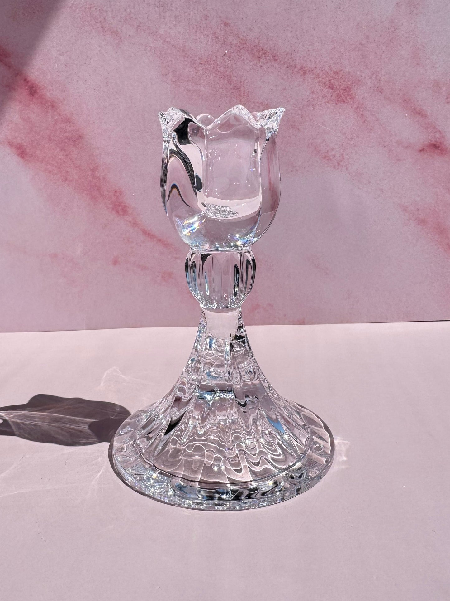 The Tulip Candle Holder, featuring clear glass with a tulip-shaped top inspired by 1990s Slovenian crystal designs, sits on a smooth pale pink surface against a pink marbled background.