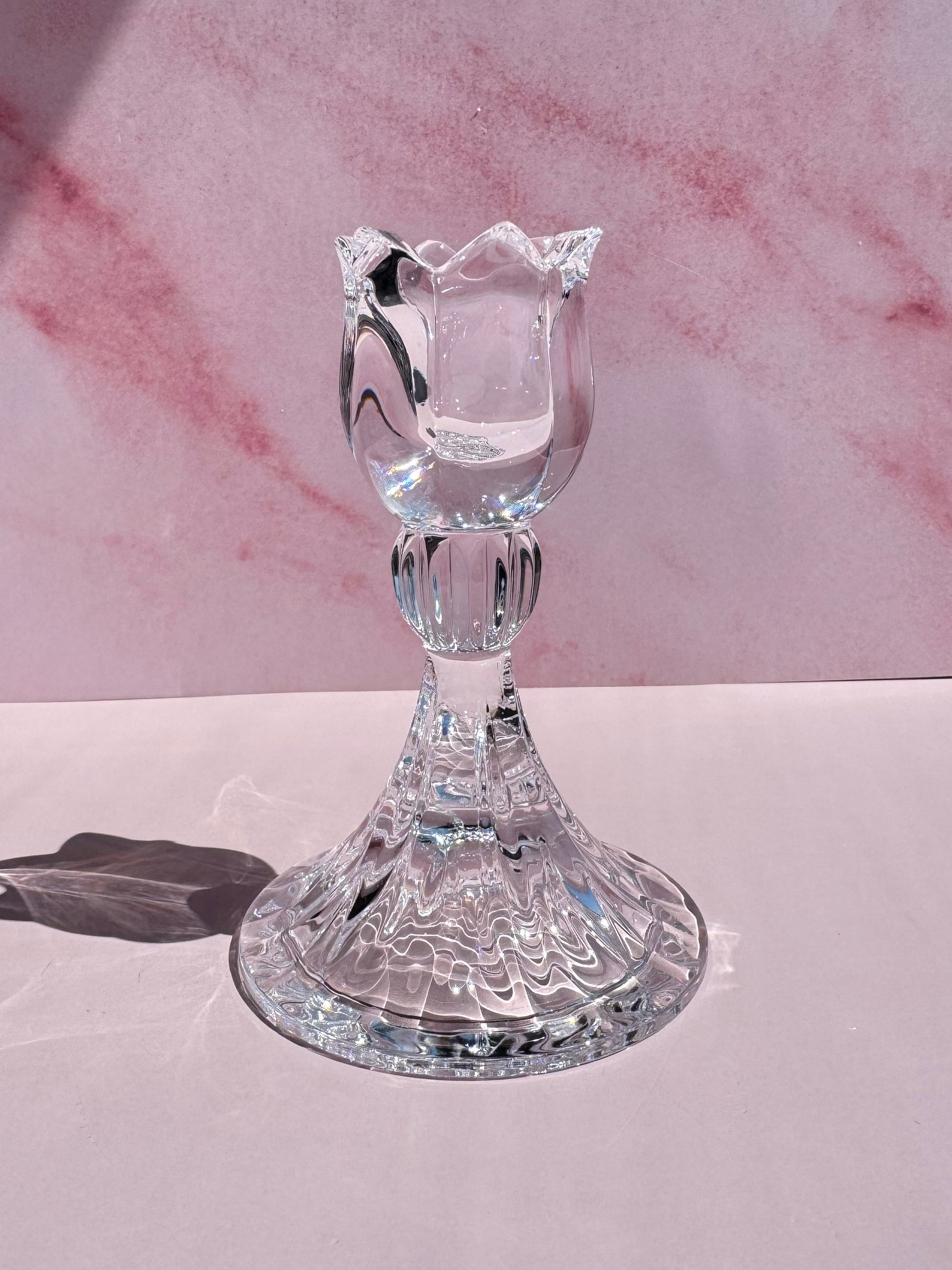 The Tulip Candle Holder, featuring clear glass with a tulip-shaped top inspired by 1990s Slovenian crystal designs, sits on a smooth pale pink surface against a pink marbled background.