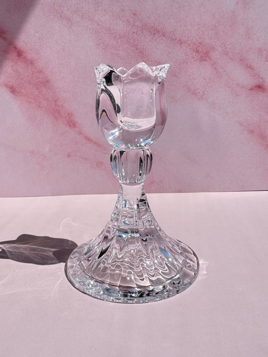 The Tulip Candle Holder, featuring clear glass with a tulip-shaped top inspired by 1990s Slovenian crystal designs, sits on a smooth pale pink surface against a pink marbled background.