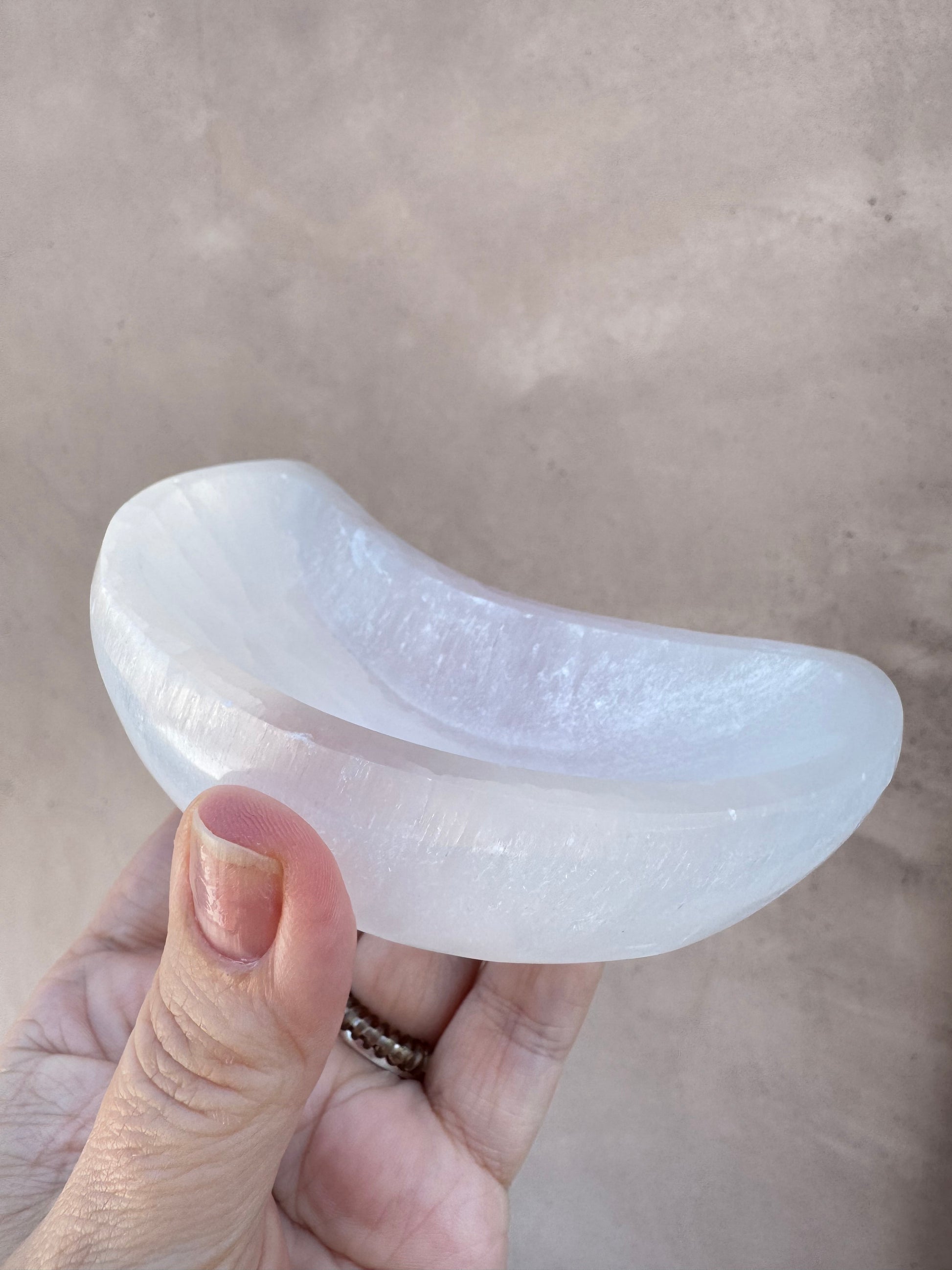 A hand holds a smooth, translucent Satin Spar Moon-Shaped Bowl, crescent-shaped and softly glowing, against a neutral background.