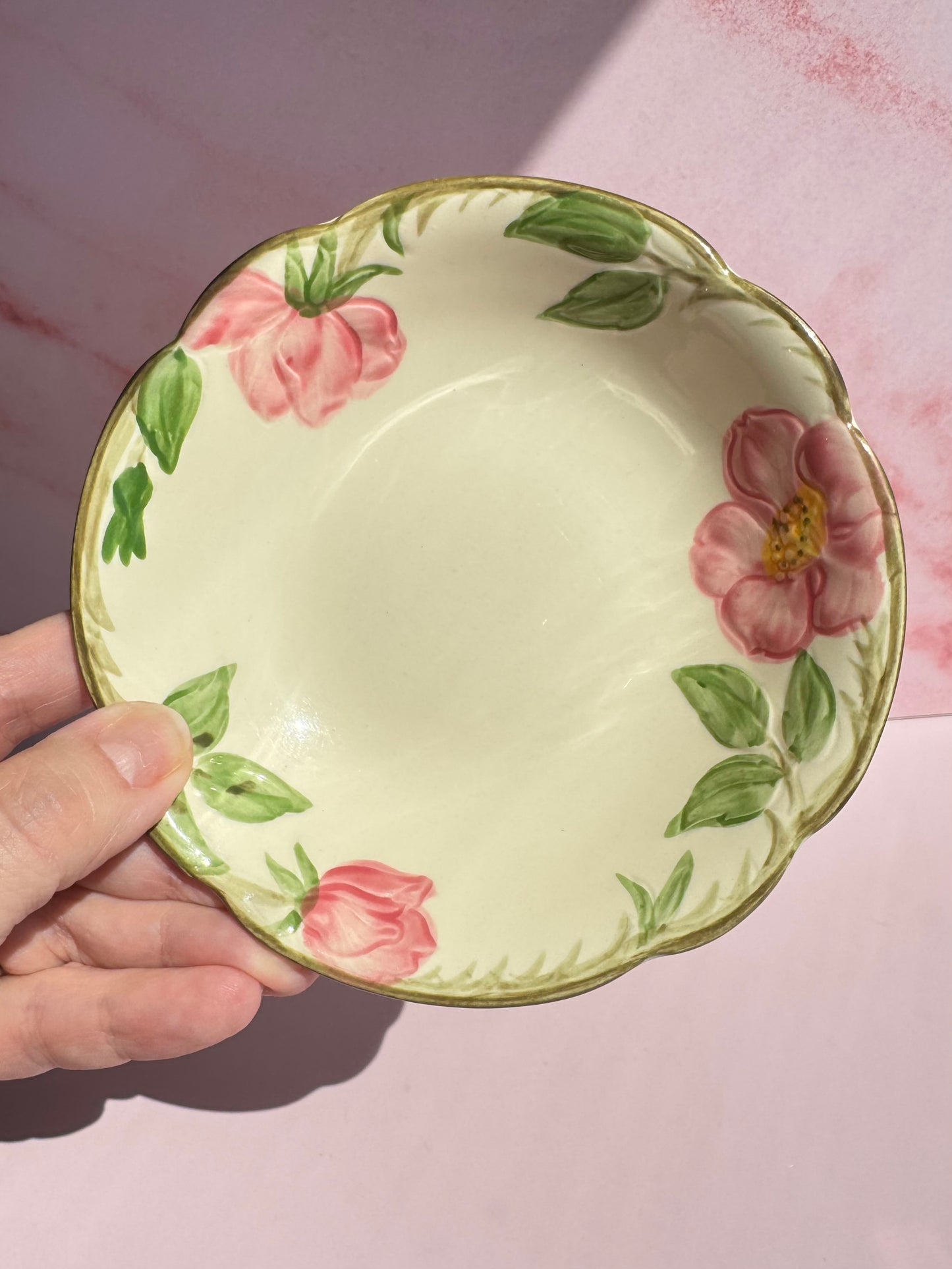 A hand holds the Desert Rose Berry Bowl, featuring a cream-colored vintage design with pink flowers and green leaves along the rim, set against a pink background.
