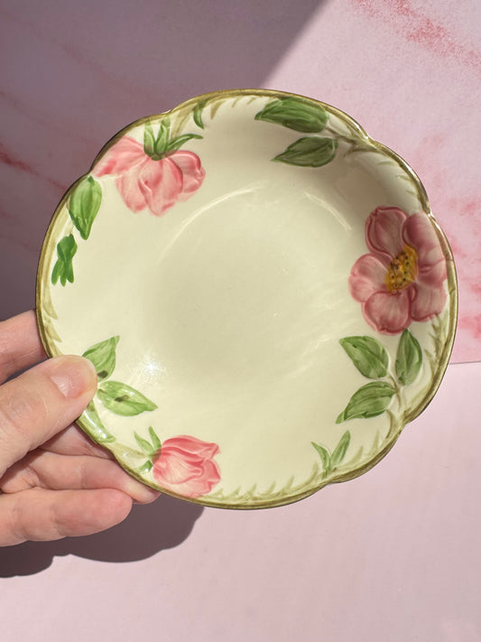 A hand holds the Desert Rose Berry Bowl, featuring a cream-colored vintage design with pink flowers and green leaves along the rim, set against a pink background.