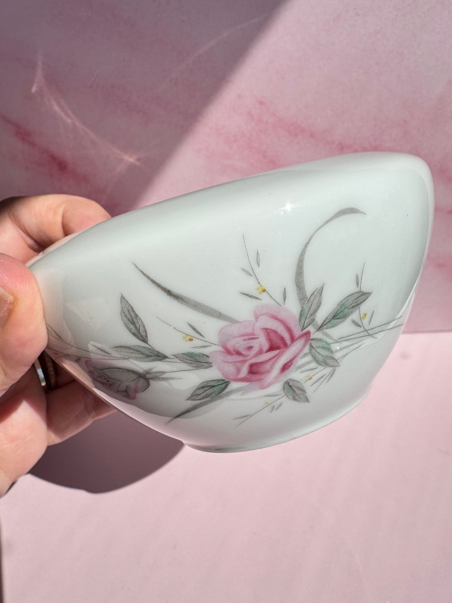 A hand holds a white Porcelain Floral Sugar Bowl adorned with a pink rose and green leaves against a pink background, evoking the charm of vintage fine china.