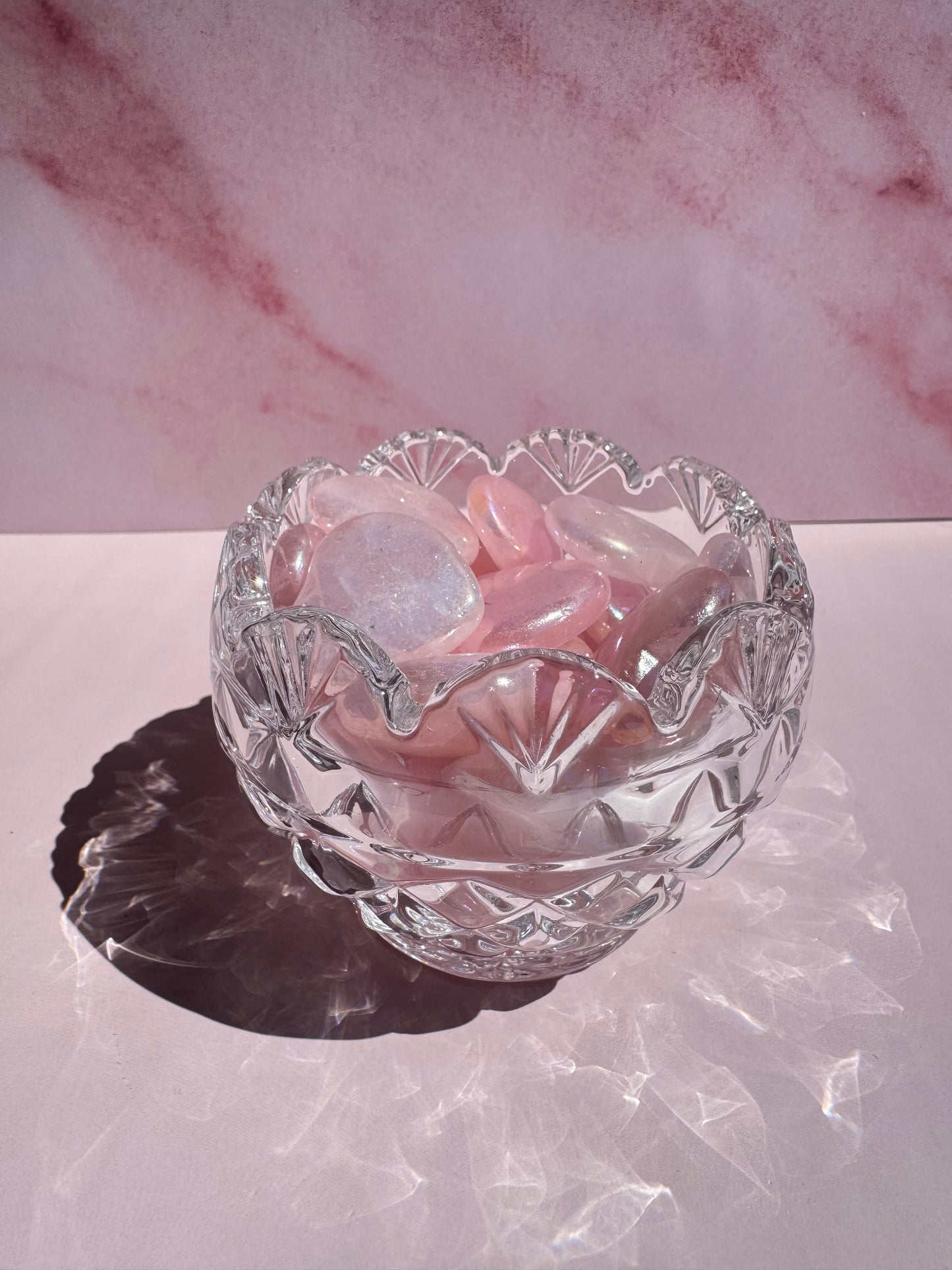 The Floral Crystal Bowl, featuring a scalloped edge and subtle flower design, displays pink and clear polished stones against a pink marbled background.
