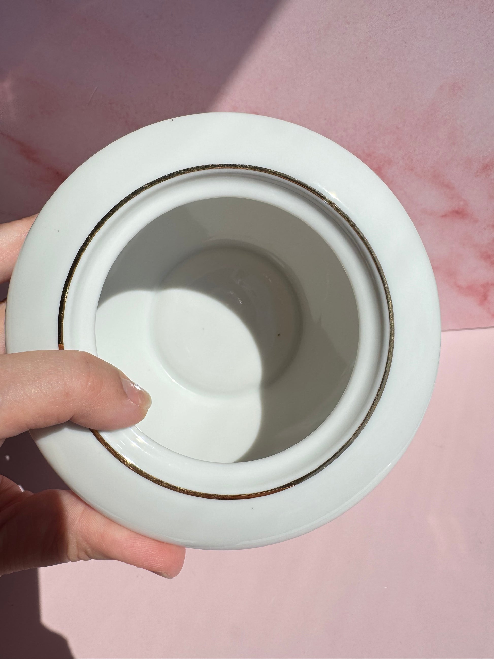 A hand holds the Porcelain Floral Sugar Bowl, its white porcelain adorned with a gold rim, poised above a pink surface where light and shadow play—a delicate piece evocative of vintage Japanese Golden Rose Fine China.
