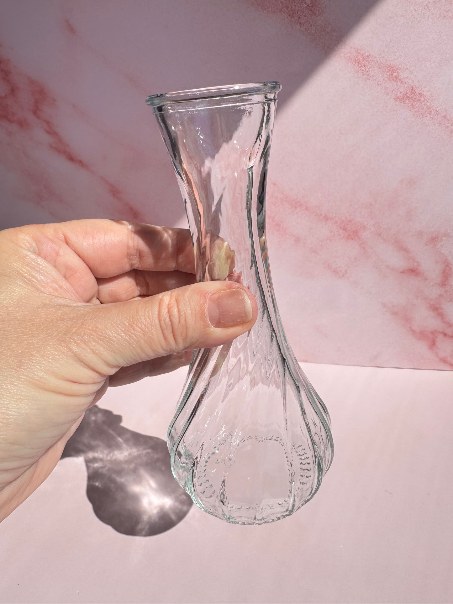 A hand holding the Glass Vase with Spiral Pattern against a pink and white marbled background in bright sunlight.