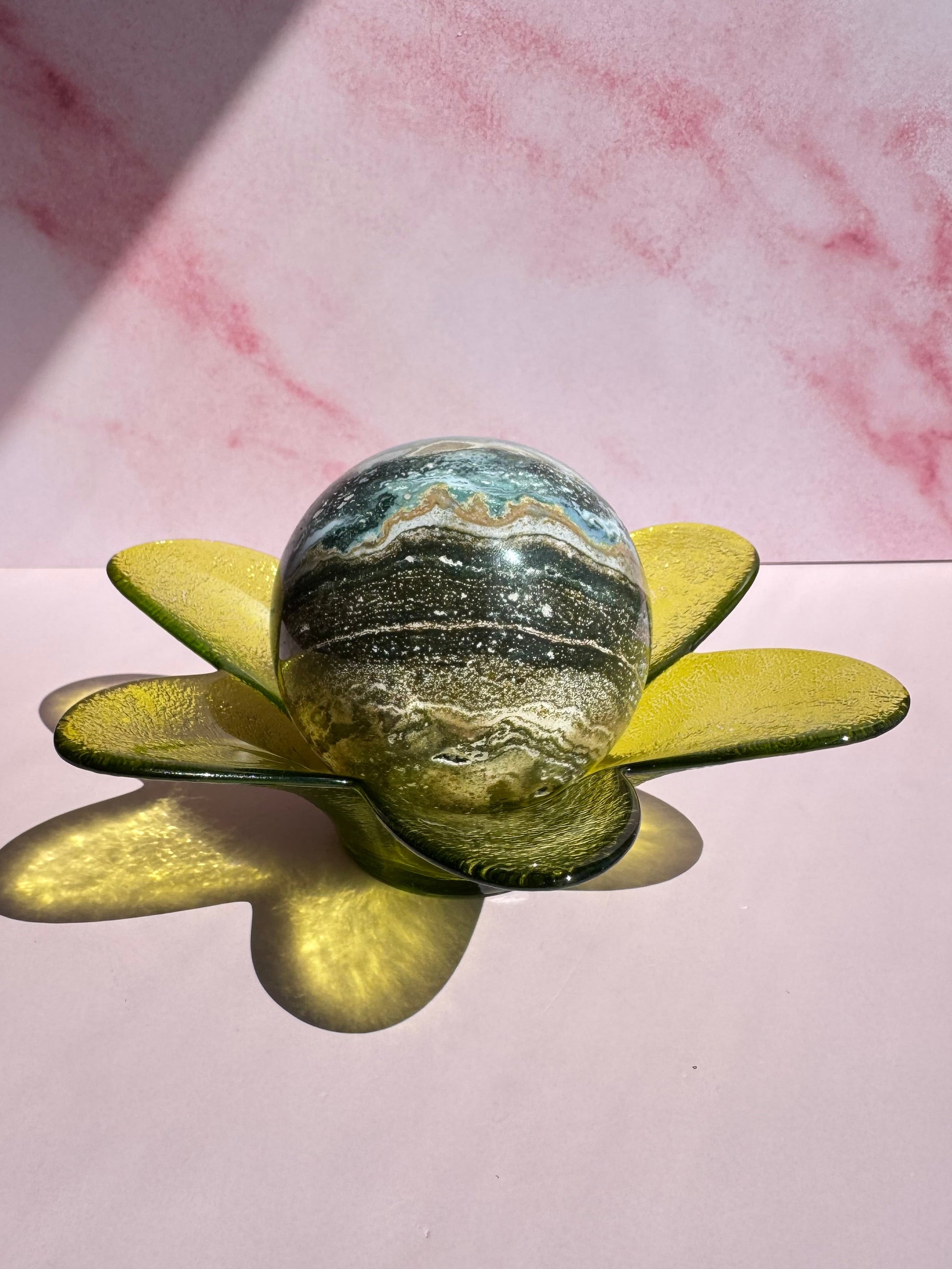 A Green Flower Glass Bowl (Imperfect) with blue, green, and brown swirls rests on a crystal holder made of yellow, petal-shaped glass pieces, casting shadows across a pink surface against a marbled pink background.