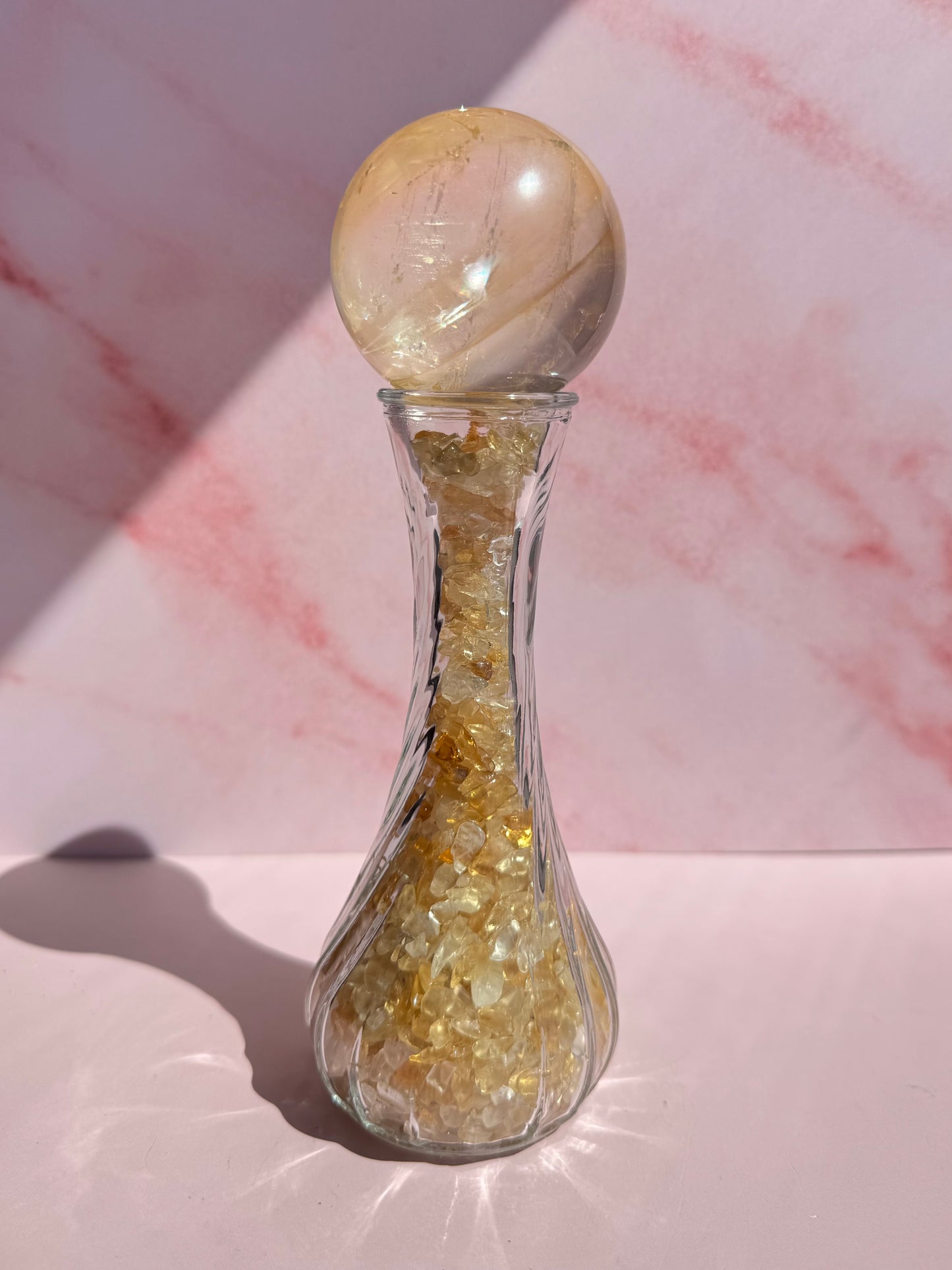 Glass Vase with Spiral Pattern, filled with yellow crystals and topped with a round crystal sphere on a sphere stand, placed on a light pink surface against a marble-patterned background.