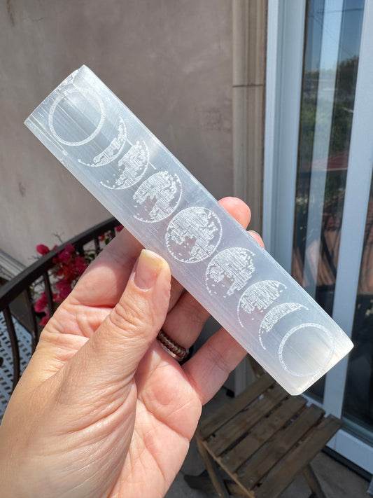A hand holds a translucent rectangular wand, identified as the Satin Spar Moon Phases Charging Bar with engraved moon phases, against an outdoor background featuring a door, window, and flowers.