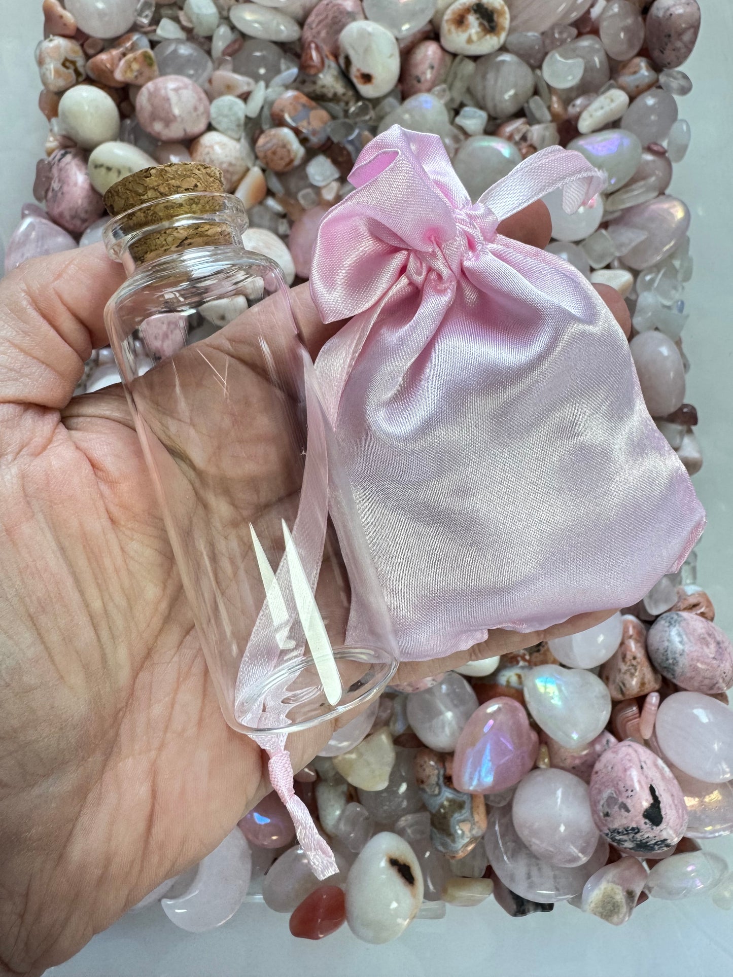 A hand holds a small empty glass bottle with a cork and a pink satin drawstring bag above polished pink and white stones from the PINK MOONGLOW MIX Crystal Confetti Scoop.