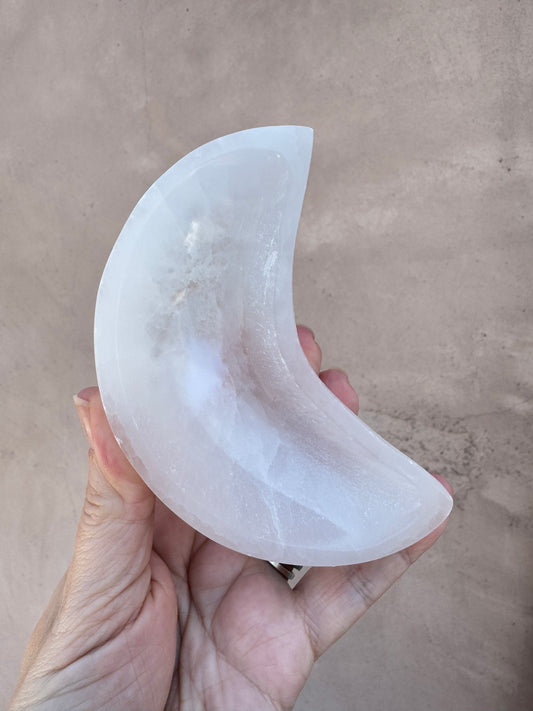 A hand holds a polished, translucent Satin Spar Moon-Shaped Bowl—its crescent form gleaming softly—against a plain, light tan background.